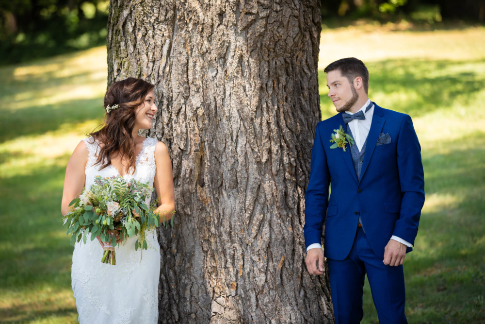 Des mariés souriants se tiennent autour d’un grand arbre. La mariée tient un bouquet et le marié porte un costume bleu.