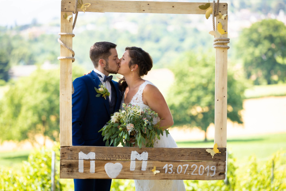 Un couple qui se marie s'embrasse dans un cadre en bois décoré, sur lequel figurent leurs initiales et la date du mariage. Ceci se déroule sur une belle scène extérieure.