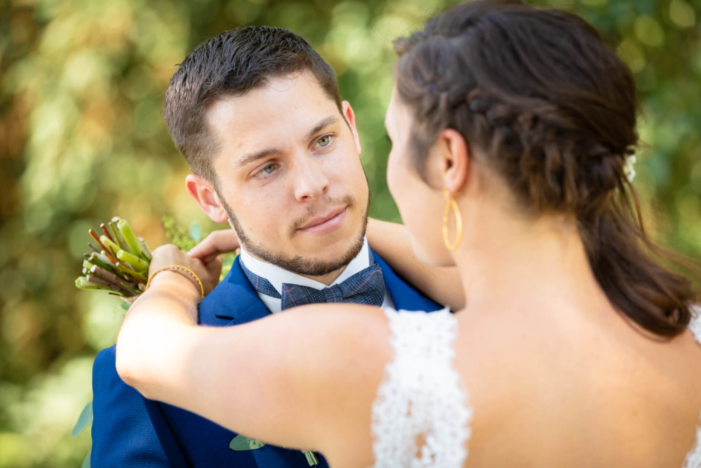 Un homme en costume bleu regarde avec tendresse sa mariée vêtue d'une robe en dentelle blanche dans un jardin baigné de soleil.