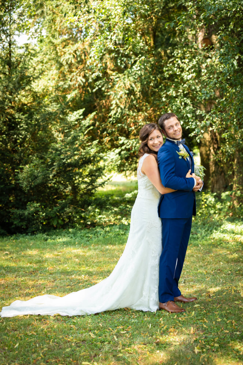 Une femme en robe de mariée blanche et un homme en costume bleu s'embrassent et sourient dans un parc ensoleillé avec beaucoup d'arbres verts derrière eux.