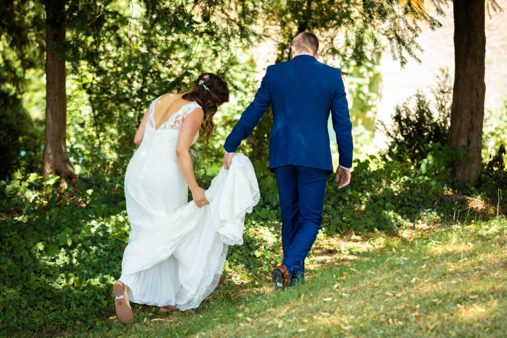 Une mariée vêtue d’une robe blanche et un marié en costume bleu se promènent main dans la main sur un chemin forestier ensoleillé. La mariée soulève légèrement sa robe en marchant.