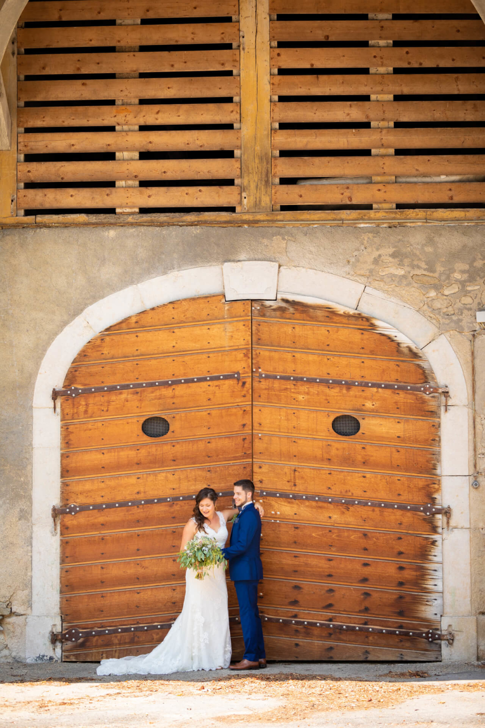Un heureux couple nouvellement marié s'embrasse devant une grande porte de grange en bois, sous une arche en pierre et un grenier en bois à lattes. La mariée a un bouquet dans les mains.