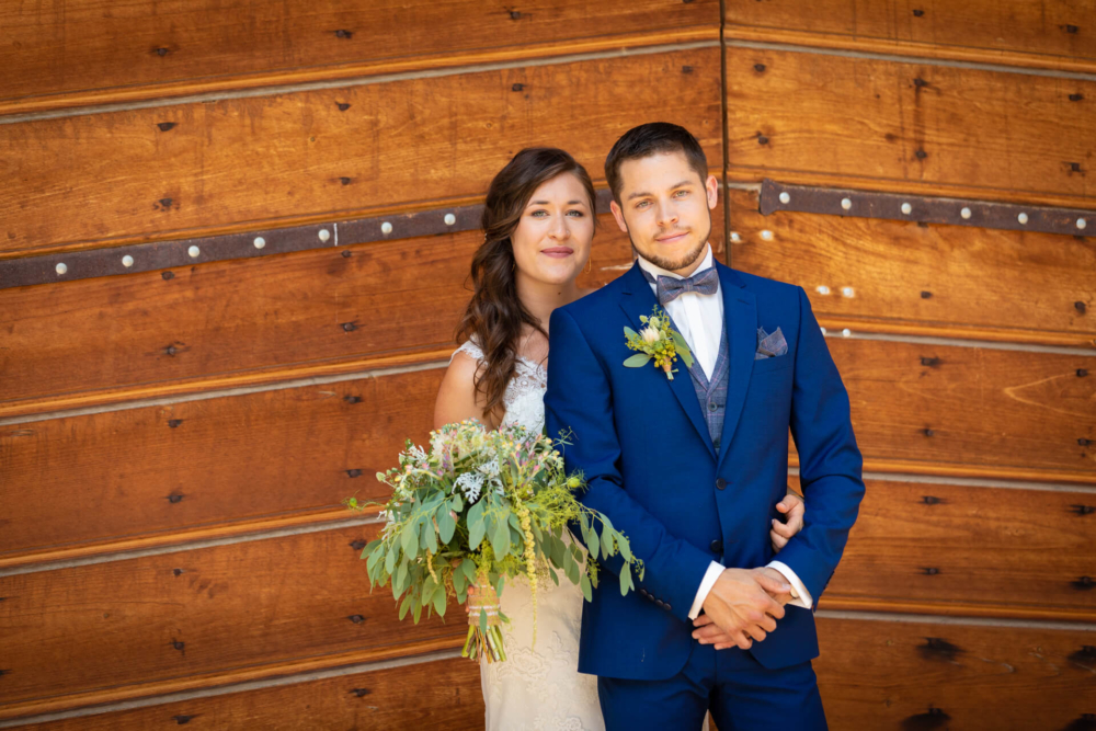 Un couple marié se tient debout et sourit doucement devant une vieille porte en bois. La mariée porte un bouquet assorti au costume bleu et à l'épingle à fleurs du marié.