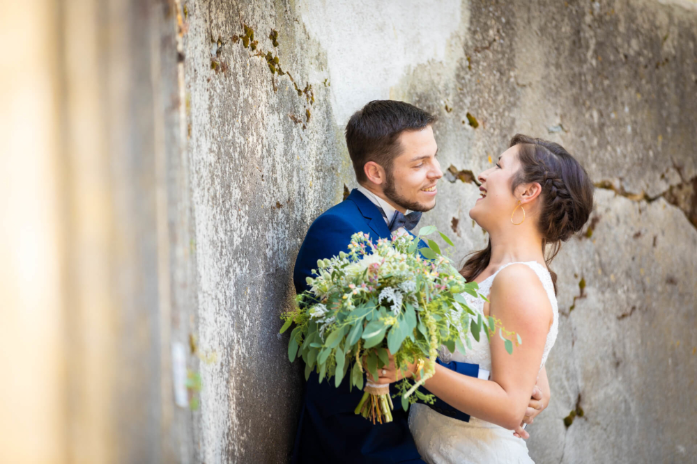 Des mariés heureux rient ensemble, appuyés sur un vieux mur. Le marié est vêtu d'un costume bleu, tandis que la mariée tient un gros bouquet de fleurs sauvages.