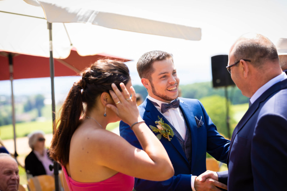 Un marié en costume bleu sourit et écoute attentivement un homme parlant lors d'une cérémonie de mariage en plein air. Une femme en robe rose joue avec ses cheveux sous le ciel ensoleillé.
