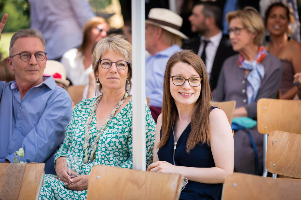 Une femme souriante vêtue d’une robe noire est assise à côté d’un couple dehors avec des gens derrière eux.