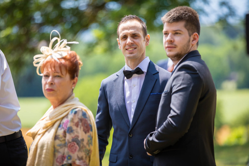 Lors d'un événement en plein air, trois invités sont attentifs. Il s’agit notamment d’un homme portant un nœud papillon qui est entouré d’une femme plus âgée portant un chapeau et d’un homme plus jeune vêtu d’un costume.