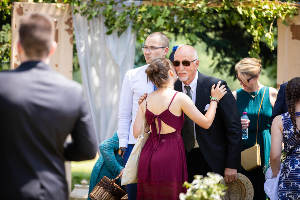 Les invités socialisent lors d’un événement en plein air. Un vieil homme portant des lunettes de soleil serre dans ses bras une jeune femme vêtue d’une robe marron. Sous le soleil, d’autres participants discutent.