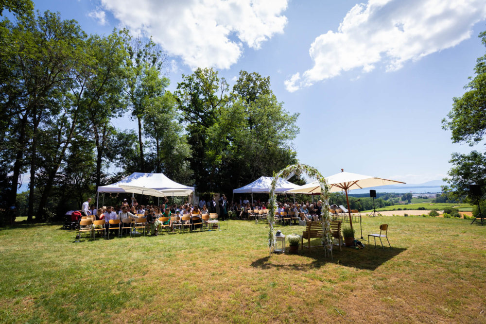 Une cérémonie de mariage a lieu en plein air, avec des invités assis sous des tentes blanches. Le temps est ensoleillé avec un ciel bleu clair. L'emplacement est entouré de nombreuses plantes vertes et d'un magnifique paysage de campagne.