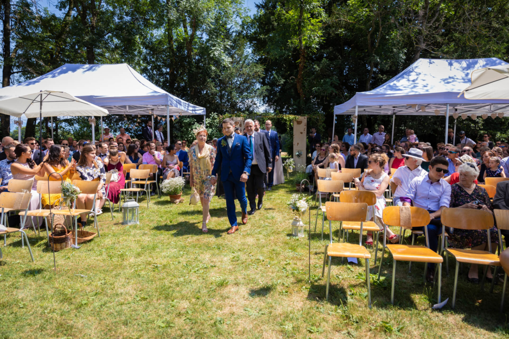 Des mariés marchent dans l’allée à l’extérieur. Ils sont entourés d'invités assis sous des parasols. C'est une journée ensoleillée et il y a des arbres en arrière-plan.