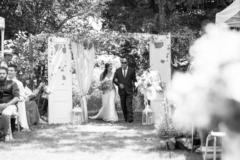 Une femme vêtue d'une longue robe de mariée blanche marche dans un chemin extérieur avec un homme plus âgé, probablement son père, lors d'une cérémonie de mariage entourée d'arches fleuries et de portes blanches.