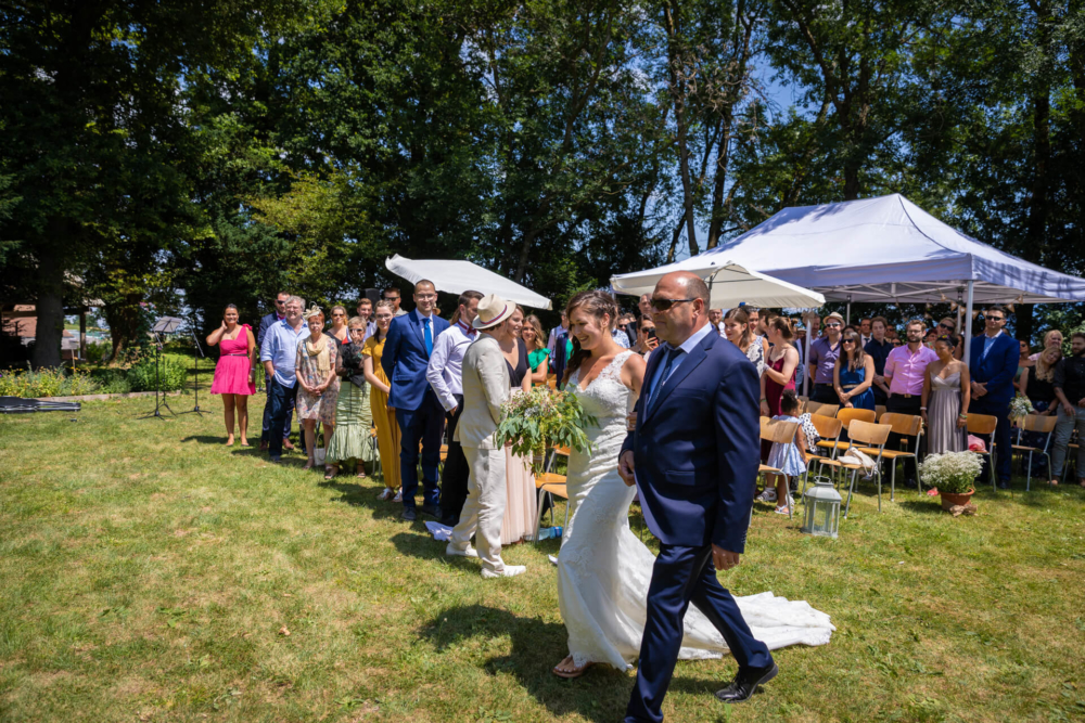Un homme plus âgé accompagne une mariée dans l'allée lors d'un mariage en plein air. Les invités sont assis et debout et regardent sous un ciel ensoleillé.