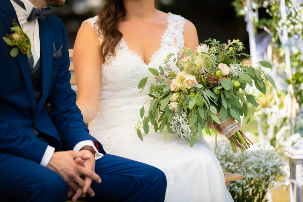 Une femme vêtue d’une robe de mariée en dentelle blanche et un homme vêtu d’un costume bleu marine sont assis l’un à côté de l’autre. La femme tient un gros bouquet de fleurs et de plantes diverses.