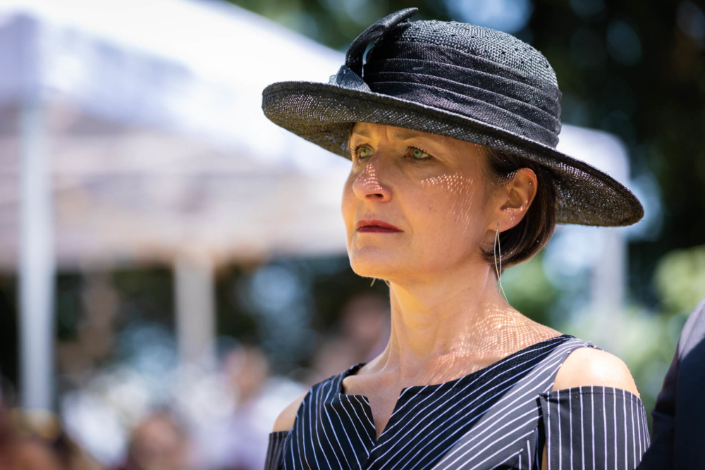 Une femme portant un chapeau noir à larges bords et une robe rayée a l'air pensive en plein soleil lors d'un événement en plein air.