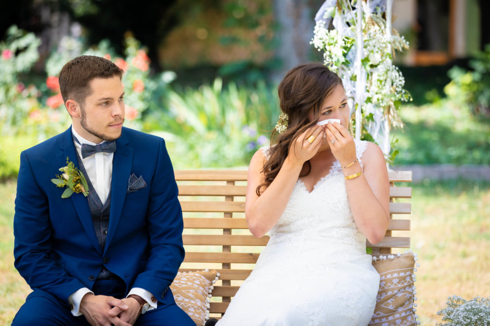 Une mariée vêtue d'une robe blanche est assise sur un banc en bois extérieur à côté d'un marié en costume bleu, tout en essuyant ses larmes. Ils sont à proximité de décorations florales.