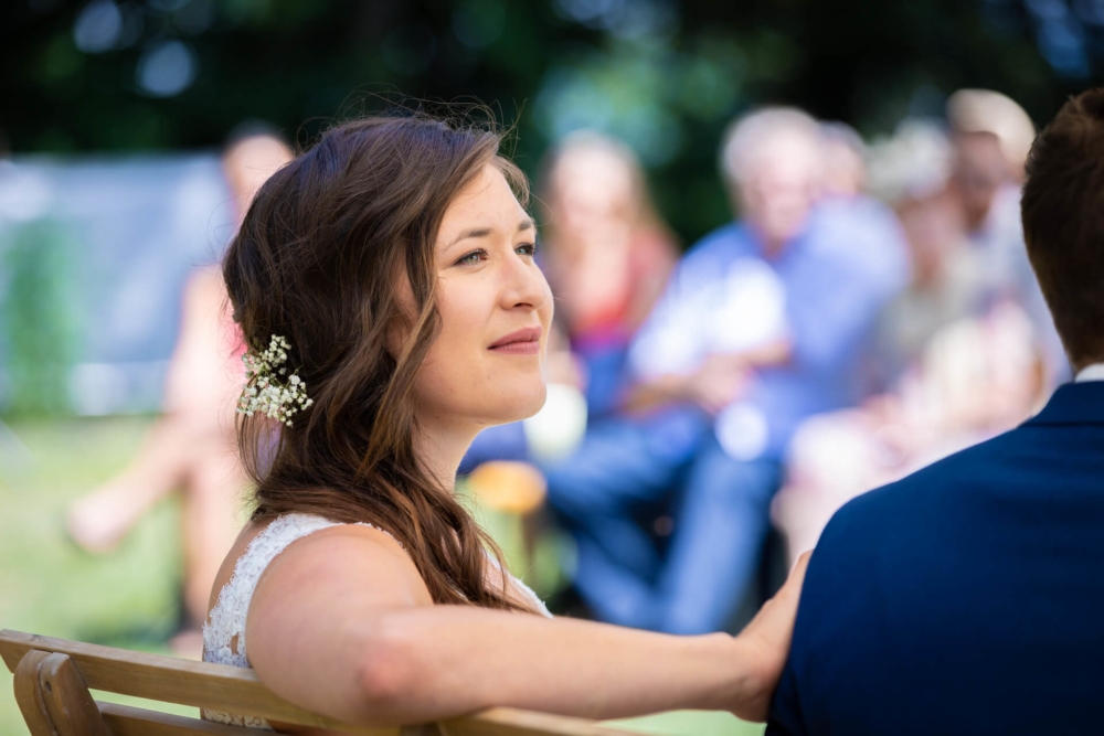 Une mariée souriante avec une fleur dans les cheveux est assise sur une chaise et regarde en arrière. Les invités au mariage sont flous en arrière-plan et le soleil brille.
