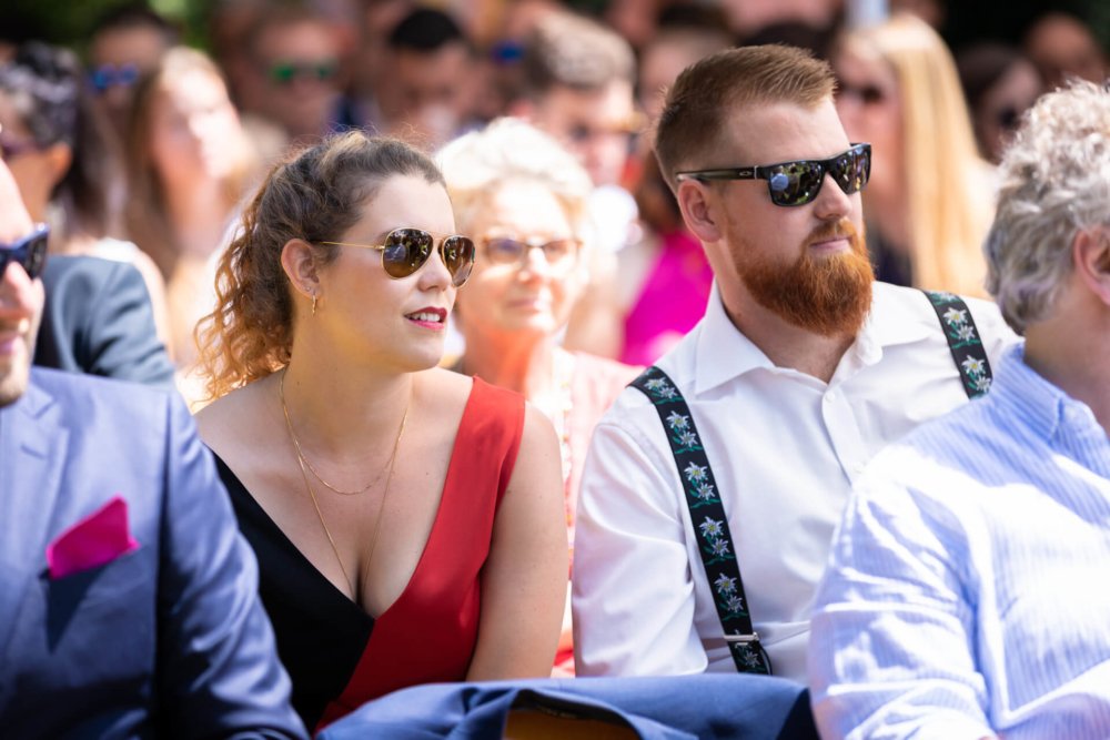 Un homme et une femme, portant des lunettes de soleil et joliment habillés, sont assis ensemble lors d'un événement en plein air. Les autres invités autour d’eux sont également concentrés sur l’événement.