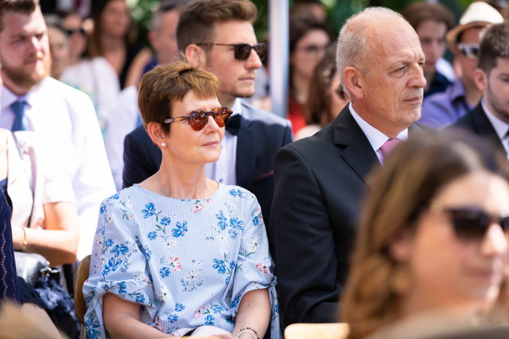 Un couple d’âge moyen surveille attentivement un événement. La femme porte une robe à fleurs et des lunettes de soleil, tandis que l'homme est vêtu d'un costume. Ils sont entourés d’autres invités également habillés de manière formelle.