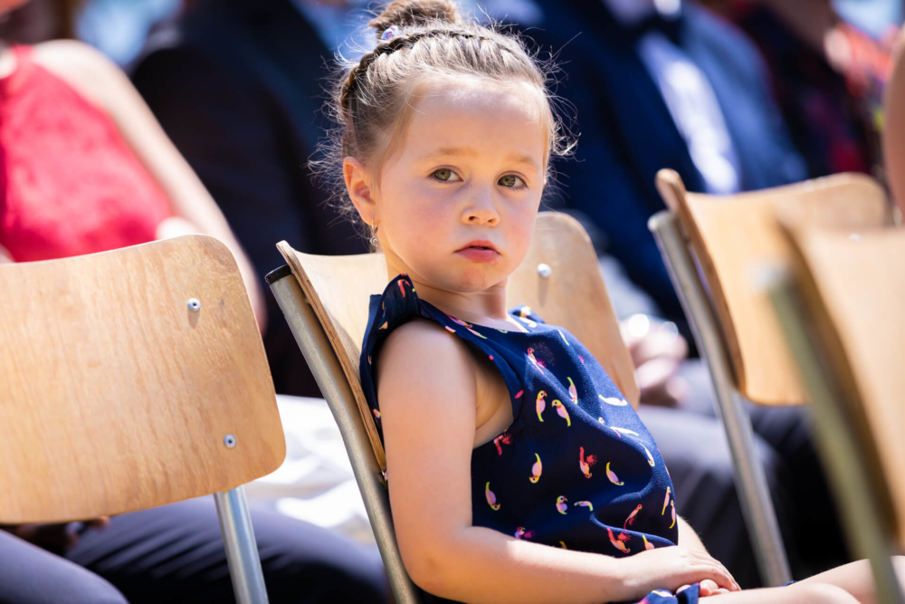 Une jeune fille en queue de cheval, vêtue d'une robe bleue à imprimé banane, est assise sur une chaise en bois lors d'un événement en plein air. Elle regarde légèrement à sa gauche avec un visage pensif.