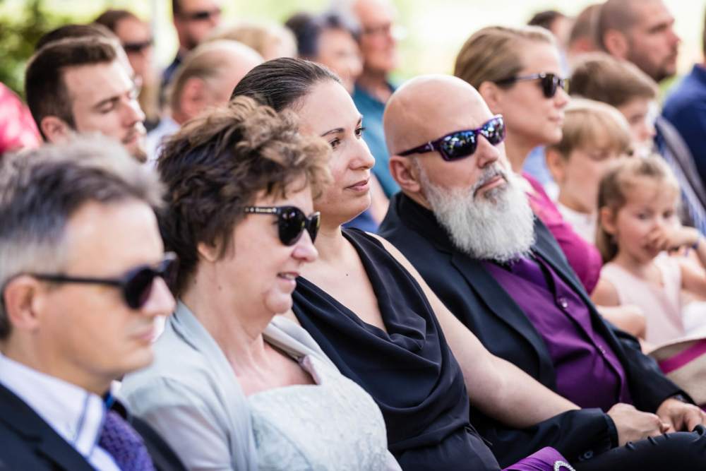 Un groupe d’adultes et d’enfants sont dehors, habillés et regardent un événement. Certains d’entre eux portent des lunettes de soleil en cette journée ensoleillée.