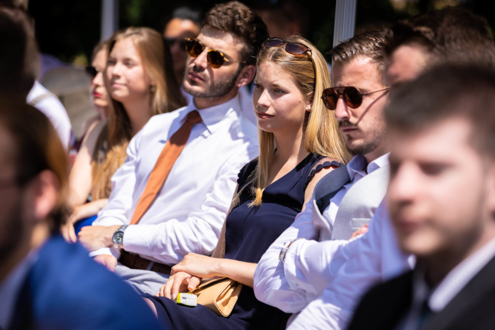 Un groupe de personnes, habillées formellement, sont assises lors d’un événement en plein air. Ils prêtent une attention particulière à quelque chose à l’avant. Avec des lunettes de soleil et des vêtements formels d’été, ils ont l’air sérieux mais intéressés.
