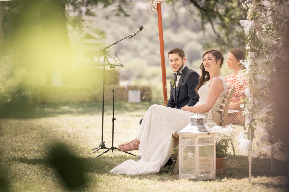 Des mariés heureux sont assis ensemble lors de leur mariage en plein air, souriant tout en écoutant un discours. L'endroit est joliment décoré avec des décorations inspirées de la nature et des lumières douces.
