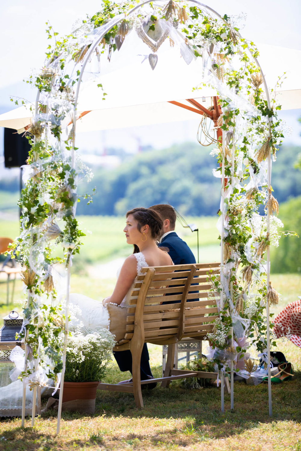 Un couple est assis sur un banc en bois sous une arche fleurie fantaisie dans un environnement de campagne ensoleillé. Ils regardent loin de la caméra vers un riche paysage verdoyant.