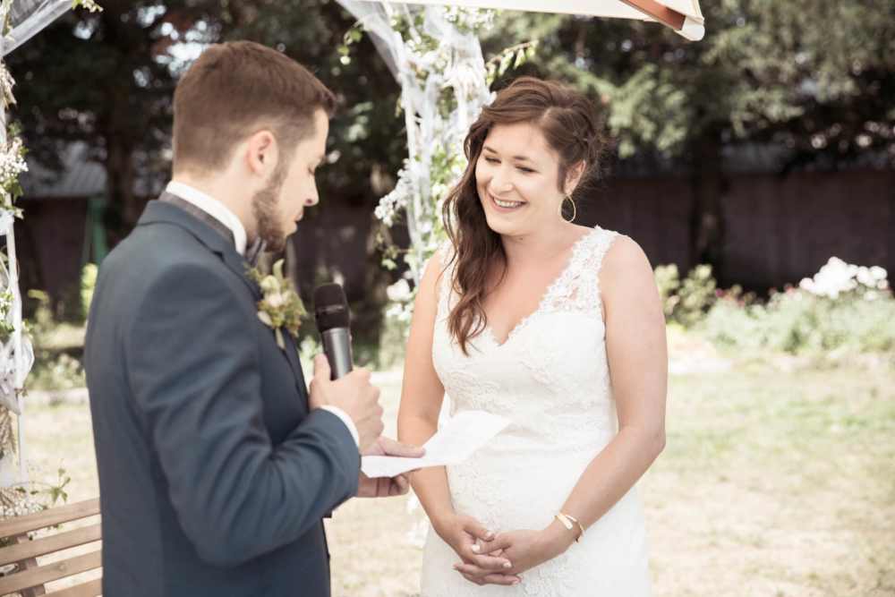 Un couple se tient sous une arche couverte de fleurs lors de leur mariage dans le jardin. Le marié lit un journal et la mariée, vêtue d'une robe en dentelle, écoute en souriant joyeusement.
