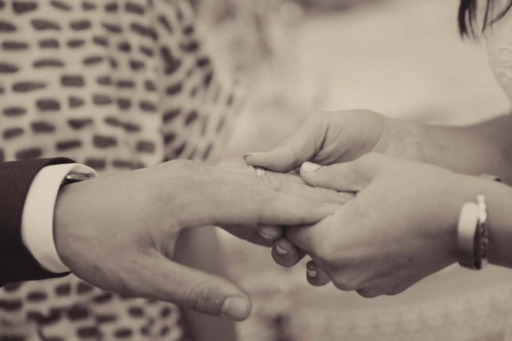Une simple photo en gros plan dans des tons brunâtres montrant une personne mettant une alliance au doigt de son partenaire, symbolisant un moment d'engagement.