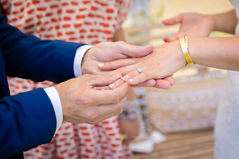 Un marié met une alliance au doigt de la mariée. Ceci est encadré par des femmes portant des robes à motifs en arrière-plan. L'attention principale se porte sur les mains et la bague, marquant un moment de fidélité.