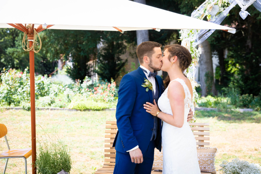 Un marié en costume bleu et une mariée en robe de dentelle blanche s'embrassent sous un parapluie blanc dans un jardin ensoleillé.