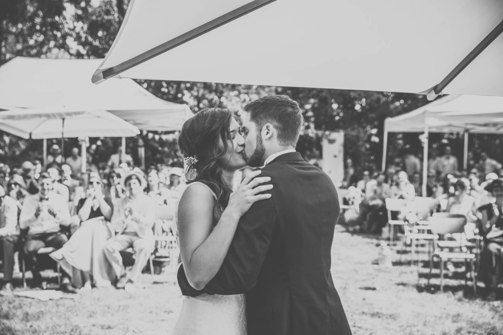 Une simple photo en noir et blanc montre des mariés s’embrassant sous une couverture. En arrière-plan, les invités sont assis dehors, applaudissant et regardant le couple.