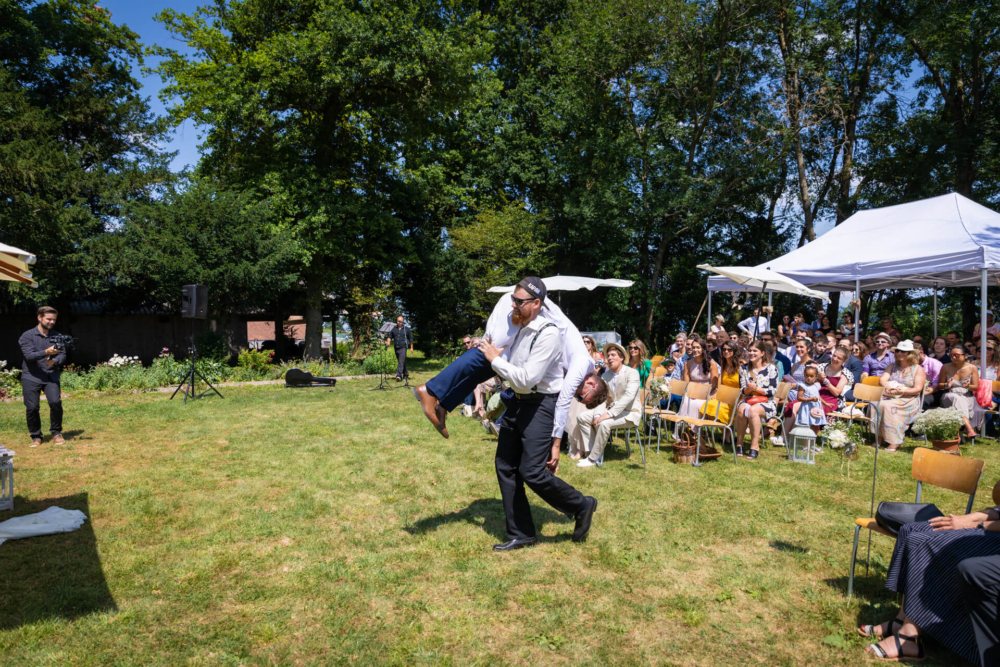 Un homme attrape joyeusement une jarretière lors d'une fête de mariage en plein air ensoleillée. Les invités sont assis sous des auvents pour regarder et profiter de la scène.