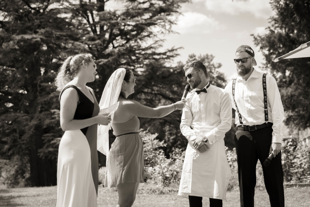 Une joyeuse photo en noir et blanc prise lors d’une cérémonie de mariage en plein air montrant les mariés riant ensemble. Une demoiselle d'honneur prononce un discours et un homme en bretelles regarde.