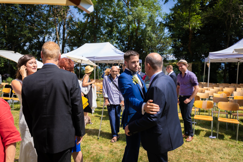 Les gens socialisent dehors lors d'un événement ensoleillé où des chaises sont installées pour une cérémonie. Certains sont installés sous des tentes blanches, le tout contribuant à une ambiance conviviale et joyeuse.