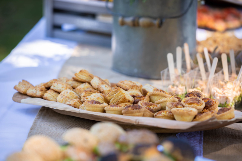 Une variété de petites tartes et pâtisseries sont présentées sur une planche de bois lors d'un rassemblement en plein air. L'arrière-plan est flou mais vous pouvez voir un seau et d'autres aliments.