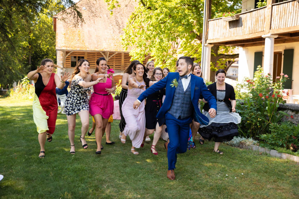 Une scène de mariage joyeuse où un marié mène les femmes dans une course amusante sous le soleil. Tout le monde sourit largement et porte des vêtements formels colorés dans un jardin luxuriant.