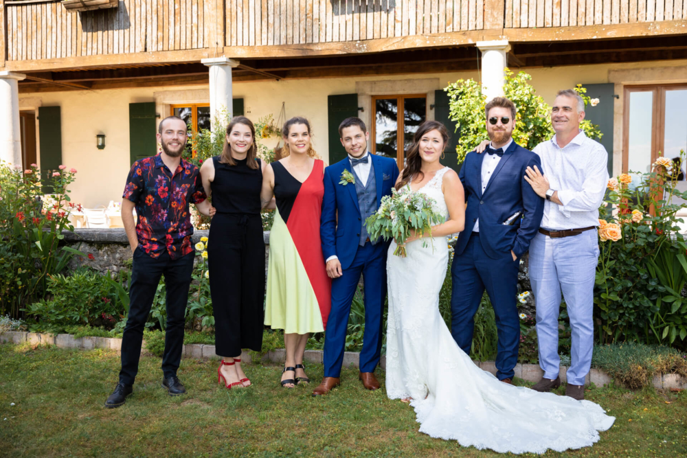 Une mariée, un marié et cinq autres personnes posent joyeusement ensemble lors d'un mariage devant une maison rustique décorée de fleurs et de plantes vertes.