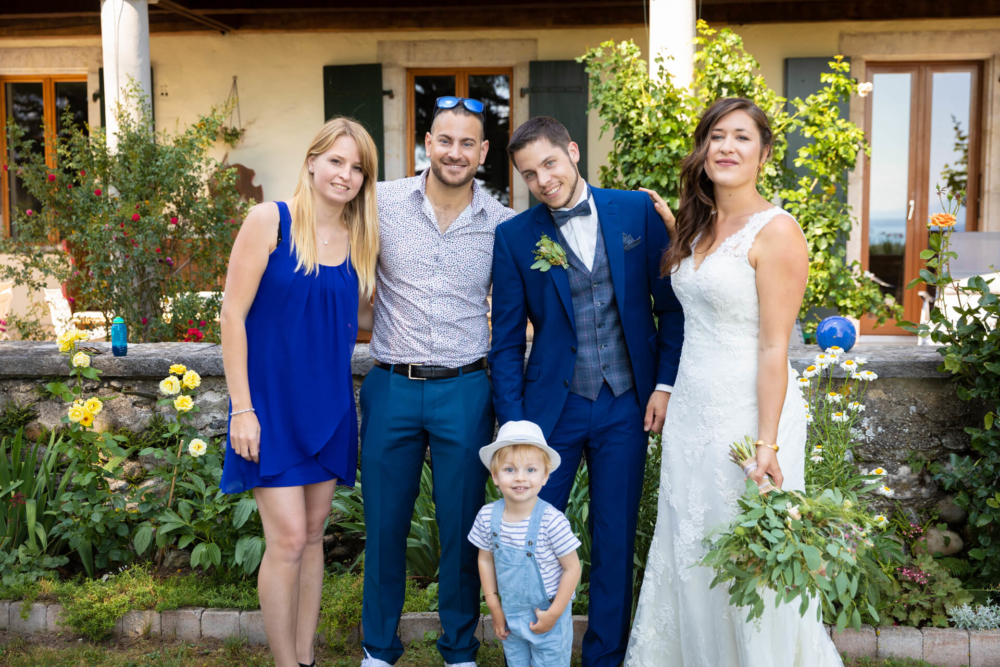Cinq personnes, dont un enfant, se réunissent lors d’un mariage devant une maison avec un jardin fleuri. Deux hommes portent des costumes et les femmes sont en robes. Une femme est la mariée qui tient un bouquet.