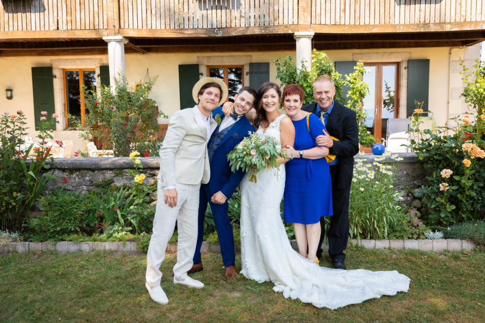 Une joyeuse fête de mariage prend une photo devant une maison de style champêtre avec un beau jardin. Les mariés sont entourés de trois invités, tous souriants.
