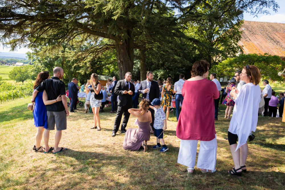 Un groupe se réunit dehors sous les arbres d’un vignoble. Ils sont en vêtements d'été, discutent et boivent un verre. Une femme devant prend une photo.
