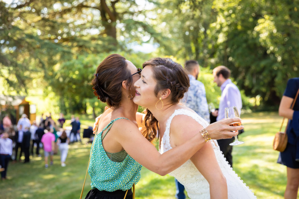 Lors d’un mariage en plein air ensoleillé, deux femmes s’enlacent et s’embrassent sur la joue. L’une porte une robe de mariée tandis que l’autre porte un haut à paillettes. Il y a des invités en arrière-plan.