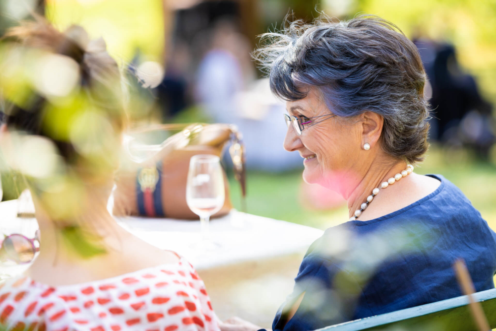 Une femme âgée portant des lunettes et des boucles d'oreilles en perles discute joyeusement lors d'un rassemblement en plein air, le dos d'une autre femme étant visible dans la scène.