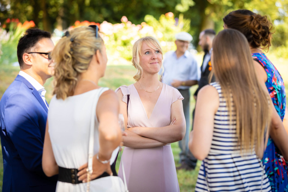 Un groupe d'adultes discutent lors d'un rassemblement à l'extérieur, y compris une dame blonde vêtue d'une robe rose qui ne regarde pas directement la caméra. Tout le monde porte des vêtements élégants et décontractés.