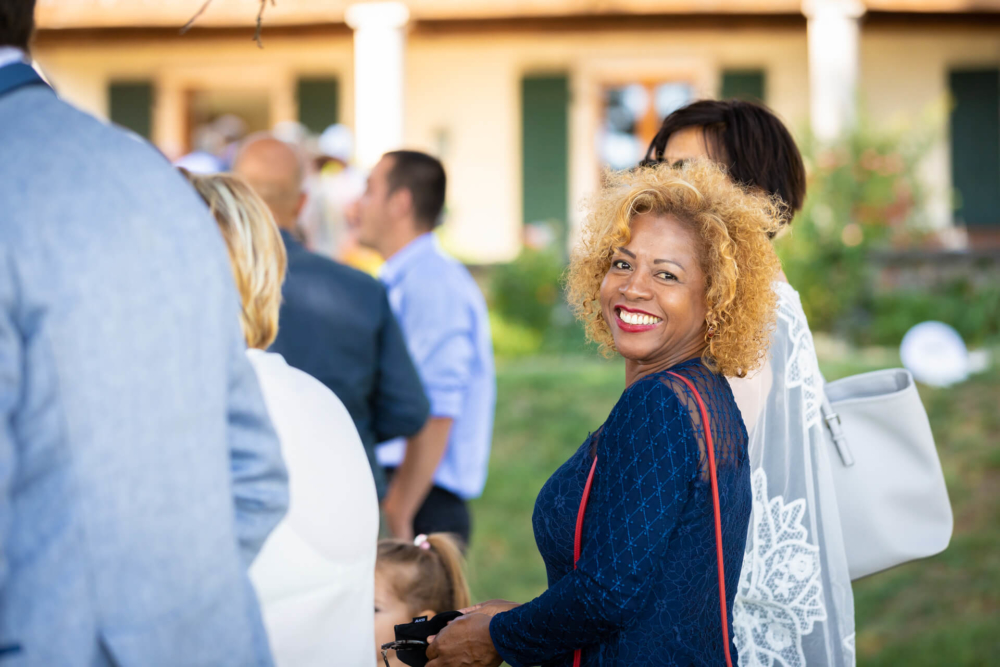 Une femme aux cheveux bouclés sourit joyeusement lors d'un mariage à l'extérieur, entourée d'invités habillés de façon formelle.