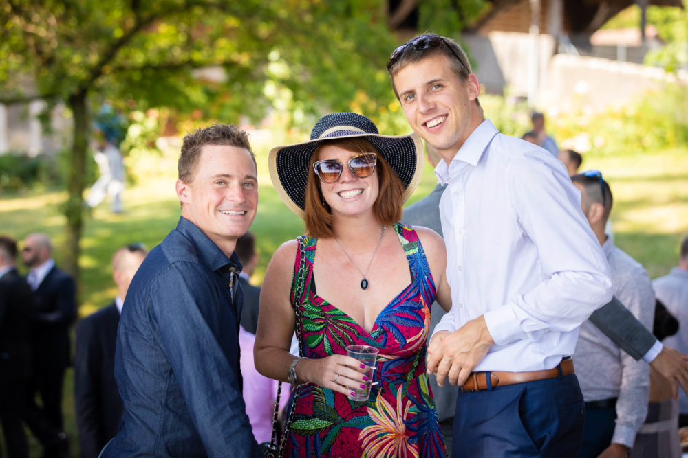 Trois adultes heureux lors d'une réunion à l'extérieur ; deux hommes et une femme du milieu vêtus d'une robe vibrante et d'un chapeau de soleil, tenant une boisson. Ils posent joyeusement pour la photo avec un parc verdoyant en toile de fond.