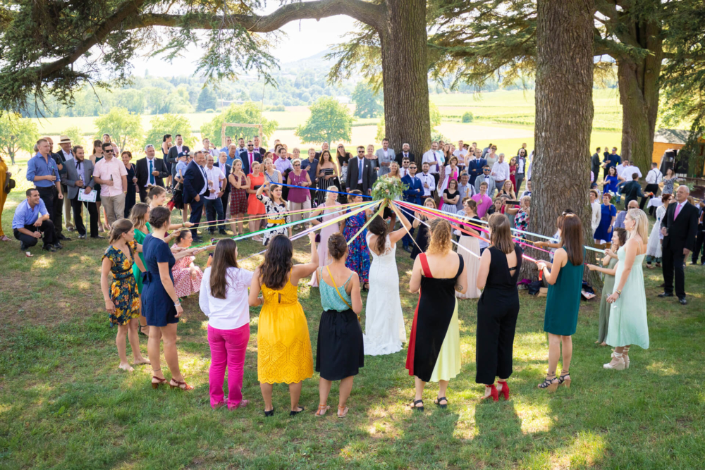 Une cérémonie de mariage en plein air où les invités entourent un couple. Les mariés se tiennent la main sous une arche, les demoiselles d'honneur portant autour d'elles des rubans multicolores.