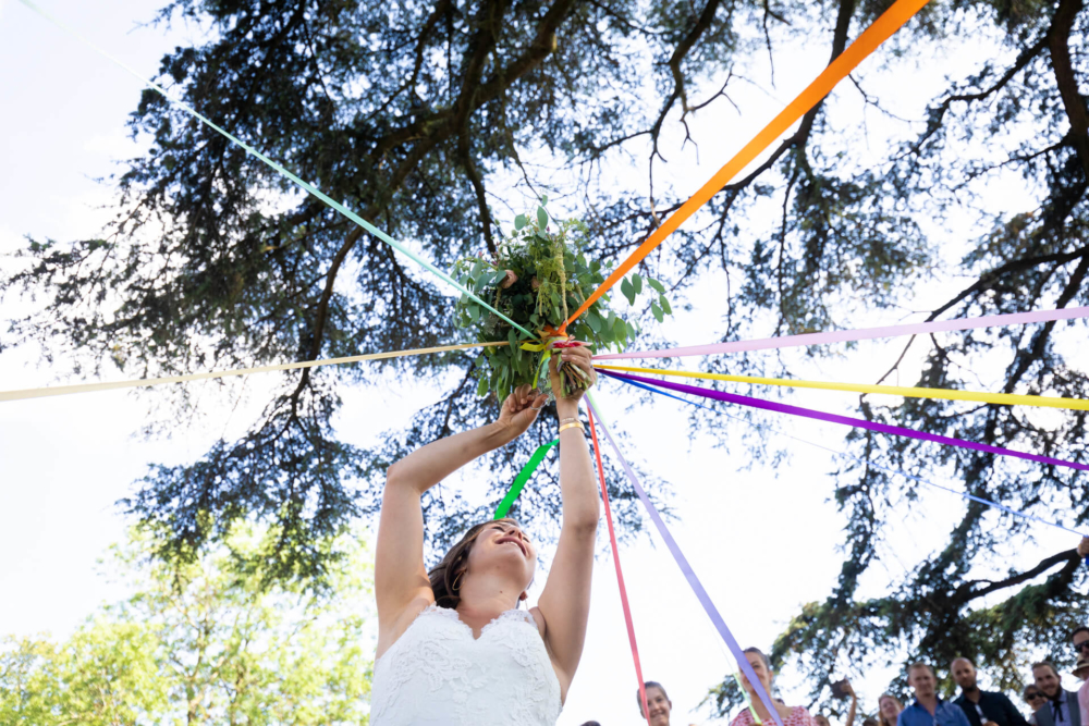 Une heureuse mariée vêtue d'une robe blanche attache des rubans sur une composition florale verte. Elle est entourée d'invités sous un décor ensoleillé rempli d'arbres.