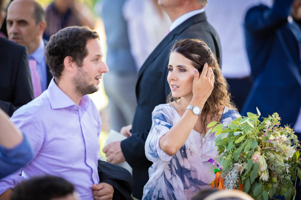 Une femme se coiffe pendant qu'elle parle à un homme. Ils sont tous les deux assis à ce qui ressemble à une fête en plein air. Elle tient un bouquet de fleurs et il y a d'autres personnes présentes à l'événement en arrière-plan.