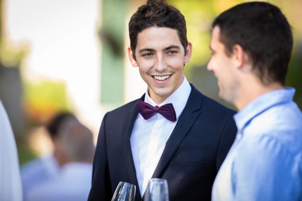 Un homme vêtu d'un smoking noir avec un nœud papillon violet, discutant et souriant avec un autre homme lors d'un rassemblement extérieur.